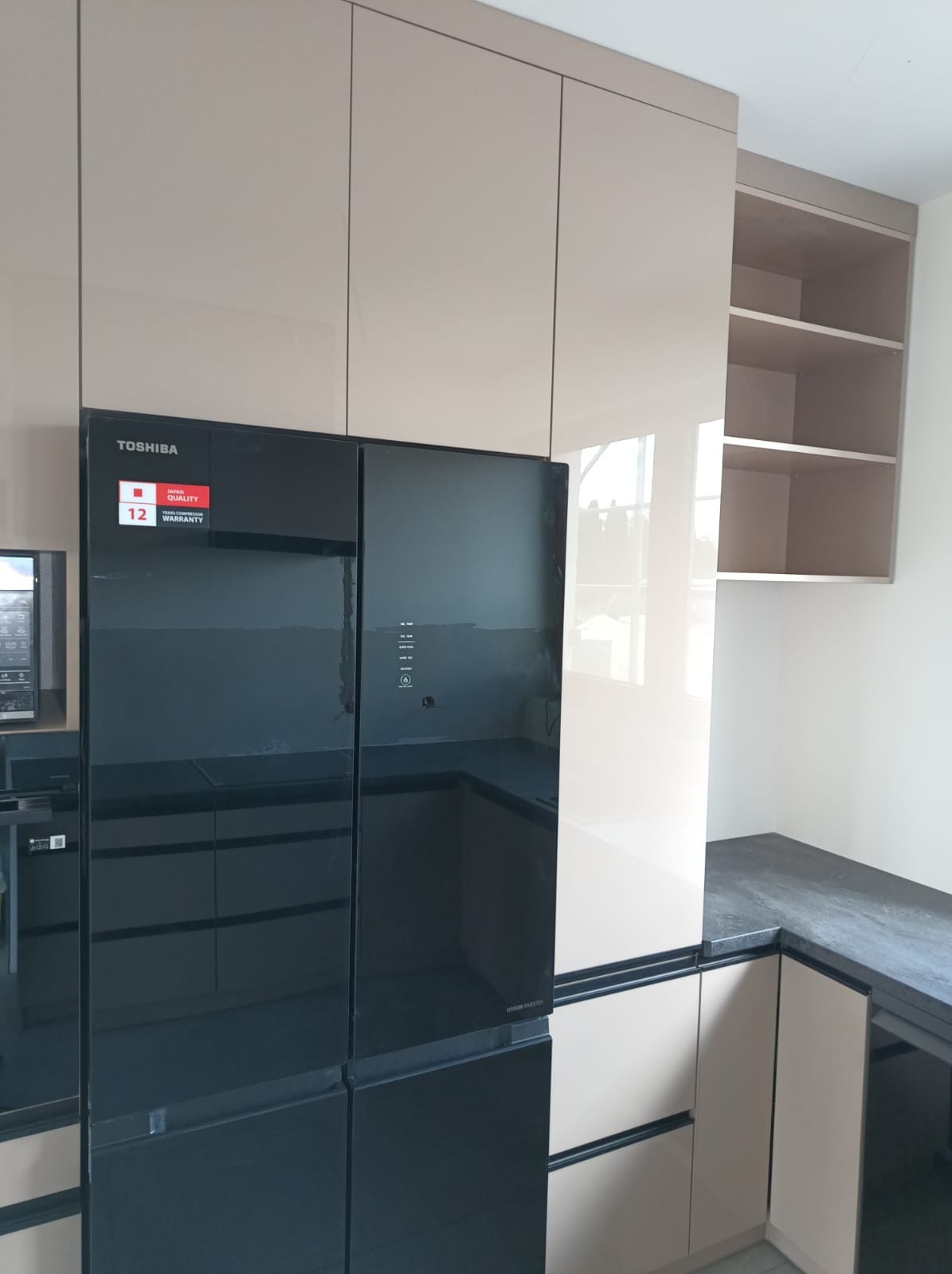 Beżowa zabudowa kuchenna z błyszczącymi frontami, czarna lodówka Toshiba z widoczną naklejką gwarancyjną, ciemny blat i otwarte półki w górnej szafce.