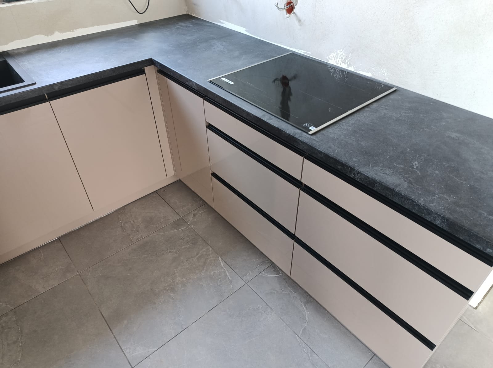 Fragment nowoczesnej kuchni z jasnymi szafkami, czarnym blatem i płytą indukcyjną, widoczne szare płytki podłogowe i niedokończone wykończenie ściany.