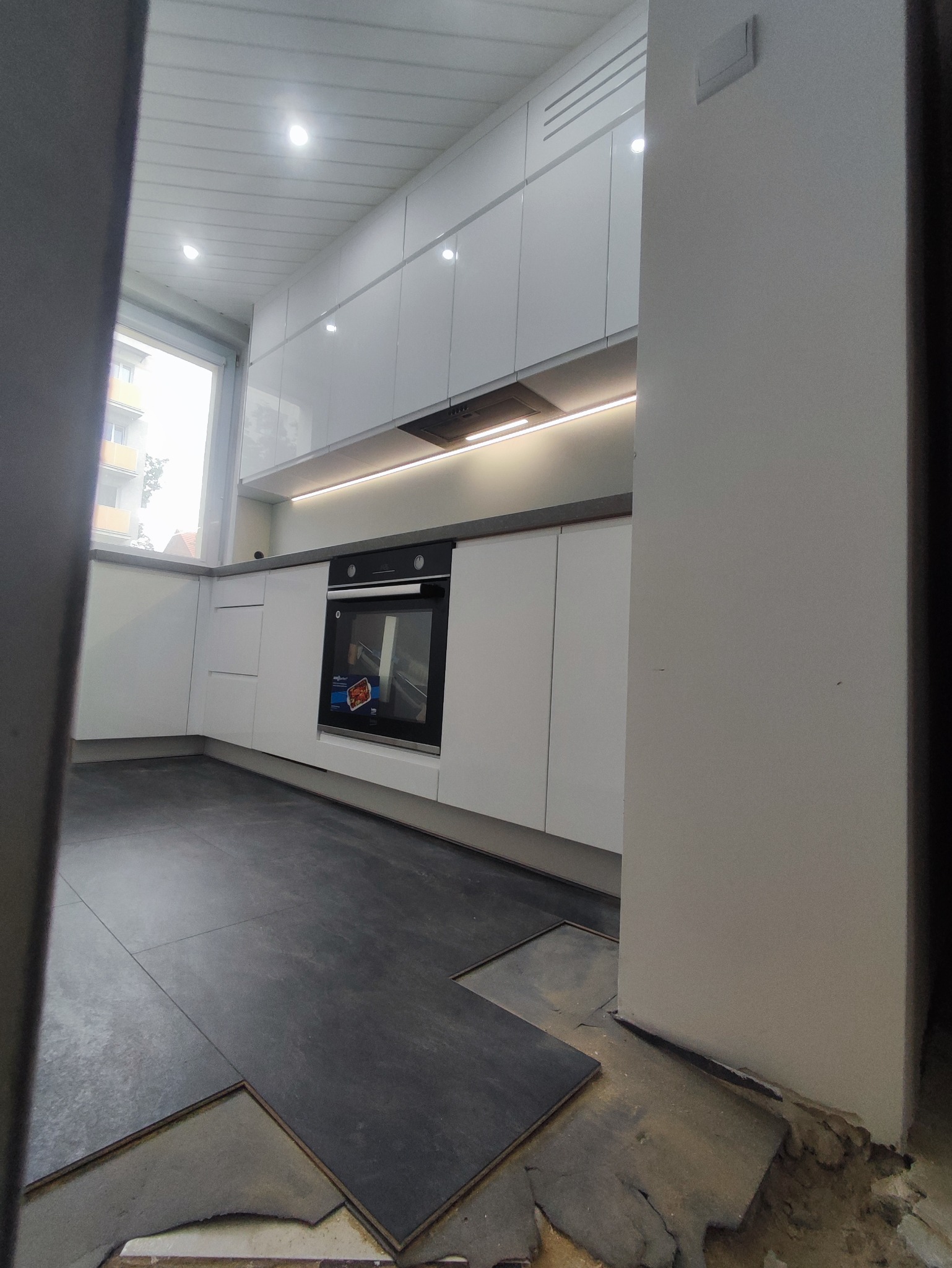 Nowoczesna, biała kuchnia z zabudową po sufit, oświetleniem LED pod szafkami i czarną płytą indukcyjną, w trakcie montażu paneli podłogowych.