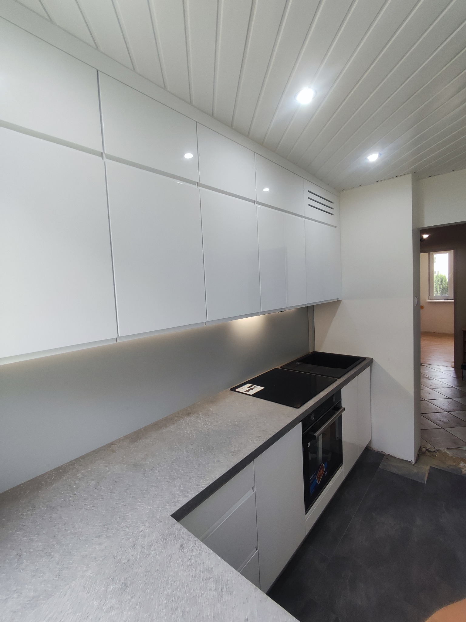 Nowoczesna kuchnia z białymi, połyskującymi szafkami górnymi z subtelnym oświetleniem LED podszafkowym, szarym blatem z wbudowaną płytą indukcyjną i piekarnikiem, oraz widoczną fragmentem podłogi...