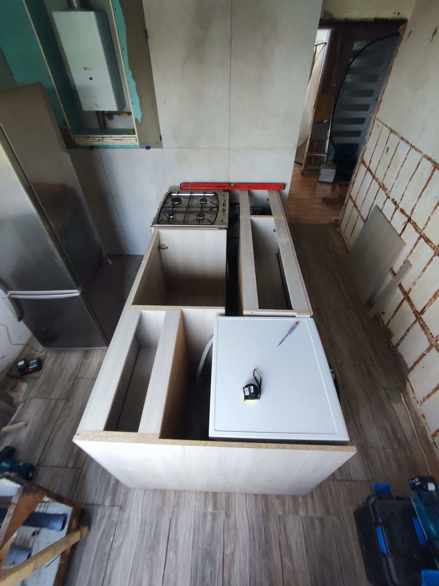 Montaż szafki kuchennej z płyt wiórowych, widoczna płyta gazowa, poziomica, narzędzia i fragmenty płytek na ścianie w trakcie remontu.