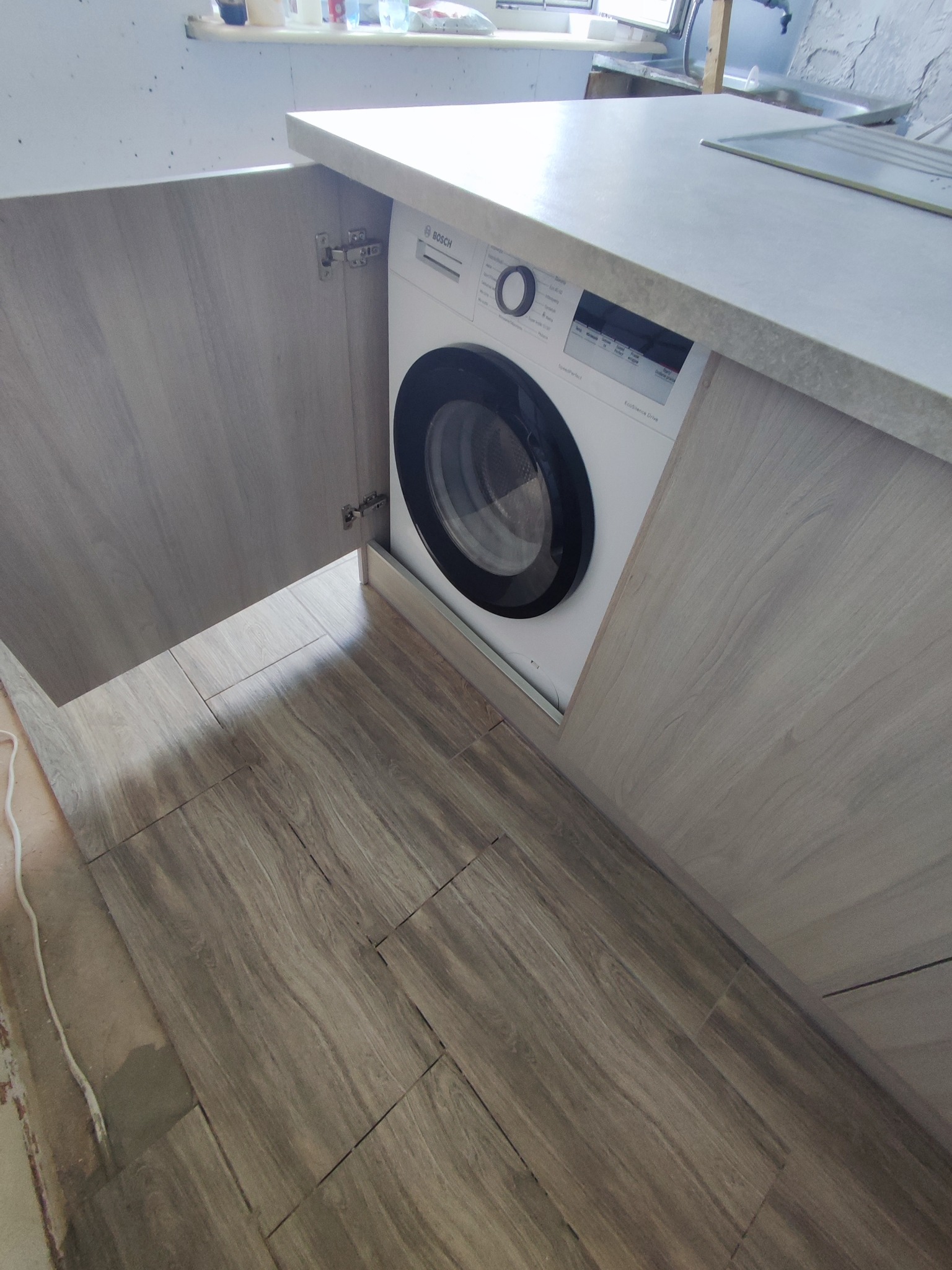 Pralka Bosch zabudowana w szarej szafce kuchennej, z blatem w kolorze betonu, widok na otwarte drzwiczki szafki i podłogę z płytek imitujących drewno.