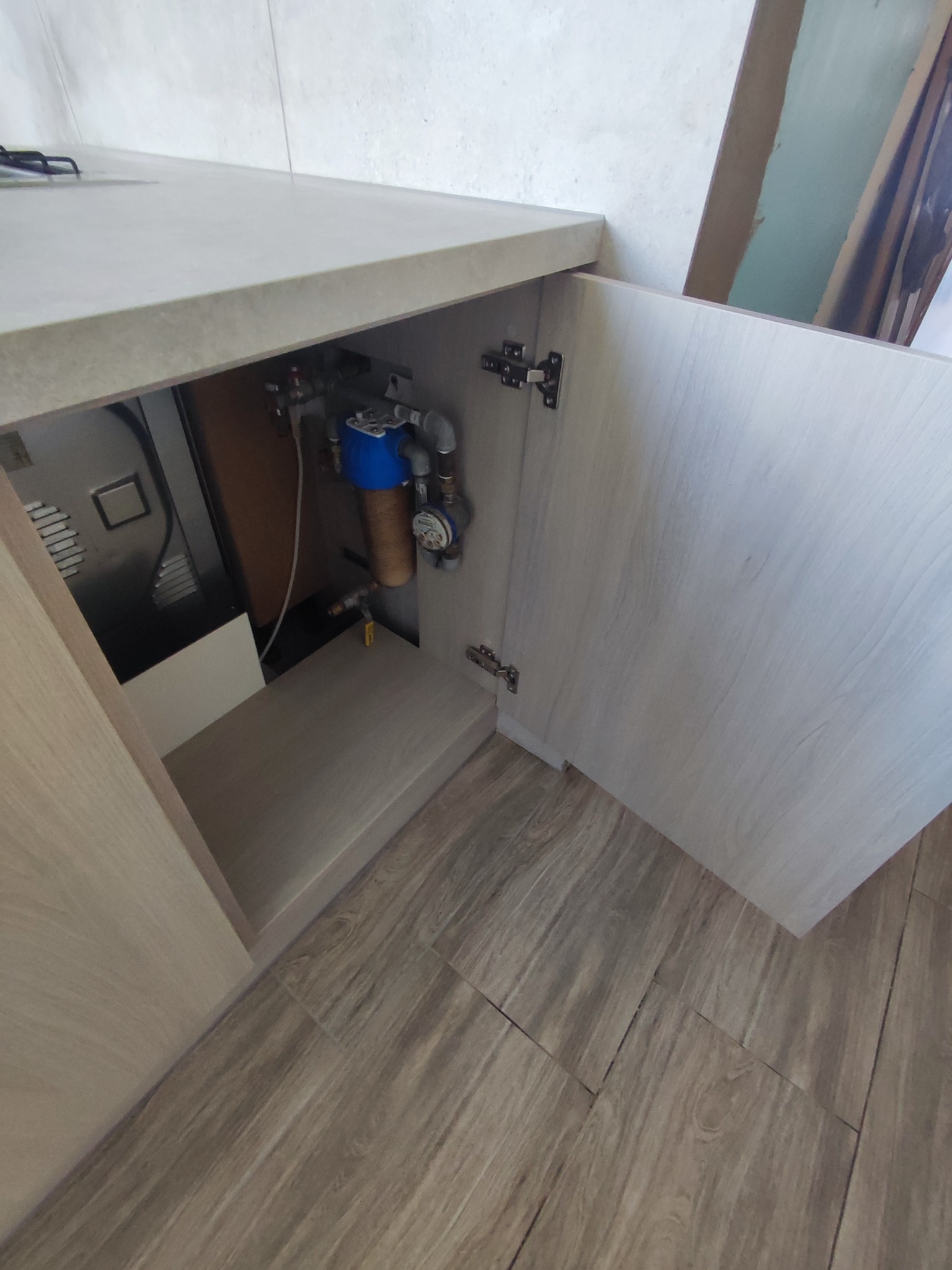 Wnętrze szafki kuchennej z filtrem wody i licznikiem, widoczne zawiasy i jasne drewnopodobne wykończenie frontów i blatu.