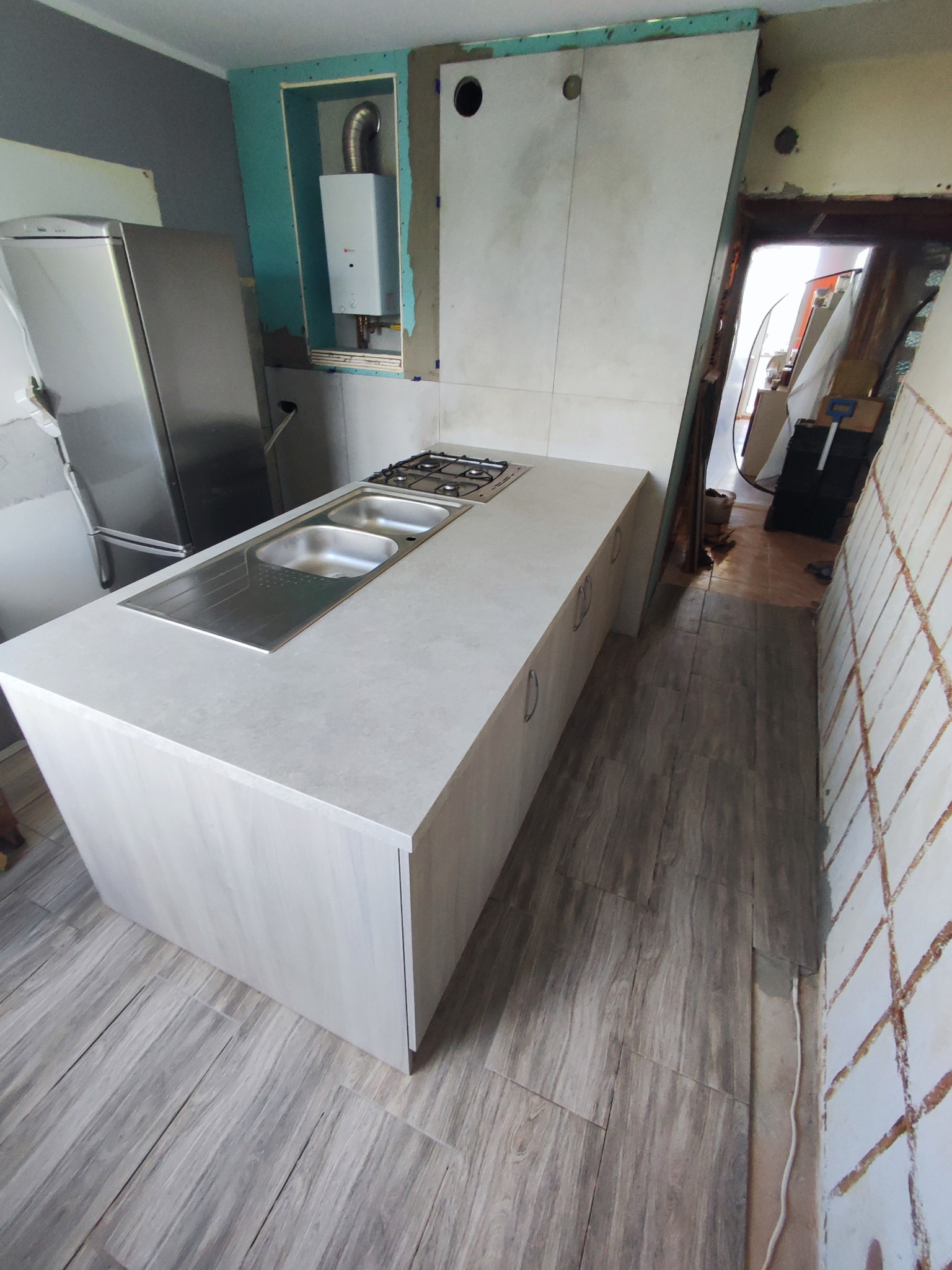 Wnętrze kuchni w trakcie remontu: wyspa kuchenna z szarą, drewnopodobną okleiną, z wbudowanym zlewem dwukomorowym i płytą gazową, na tle surowych ścian i nowego bojlera.