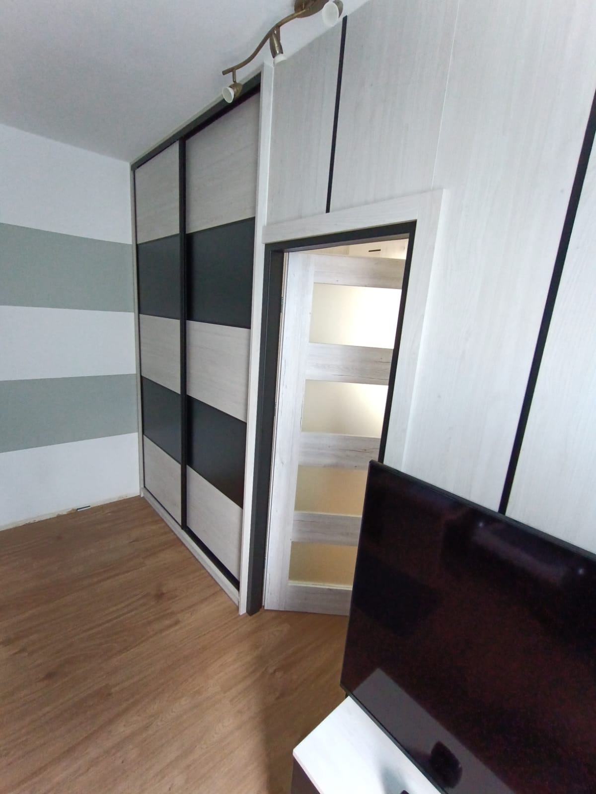 Szafa wnękowa z przesuwnymi drzwiami w kolorze jasnego drewna i czerni, widoczna z boku, z fragmentem telewizora w kadrze, na podłodze panele drewniane.