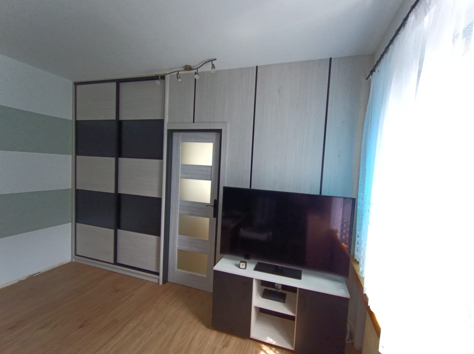 Nowoczesna szafa przesuwna z czarno-beżowymi panelami, dopasowana do ściany z drewnianymi panelami, obok drzwi z przeszkleniami, w pokoju z drewnianą podłogą i telewizorem.