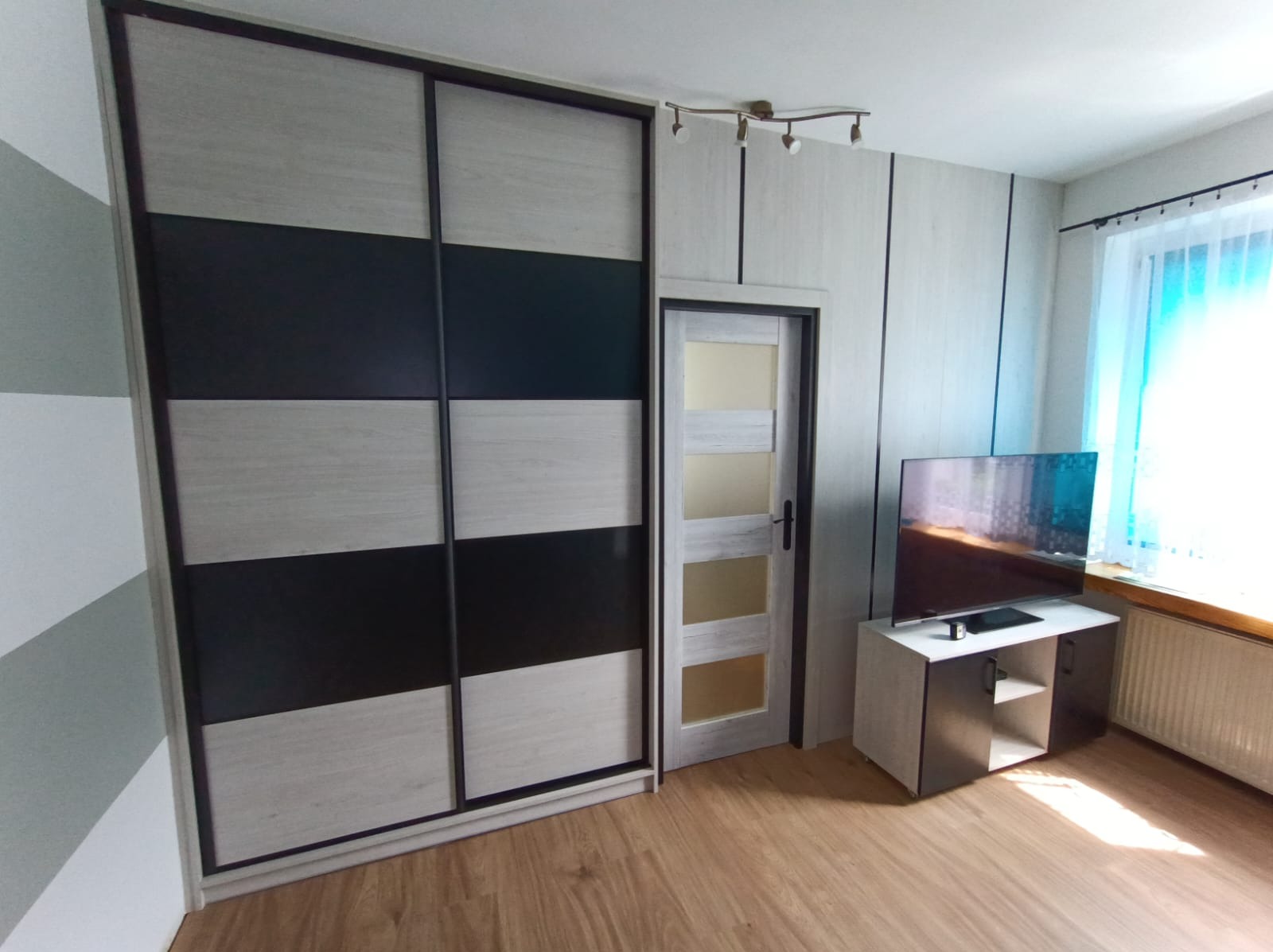 Szafa przesuwna z frontami w poziome pasy czerni i jasnego drewna, dopasowana do drewnopodobnej okleiny na ścianie i drzwiach, w tle okno z firanką.