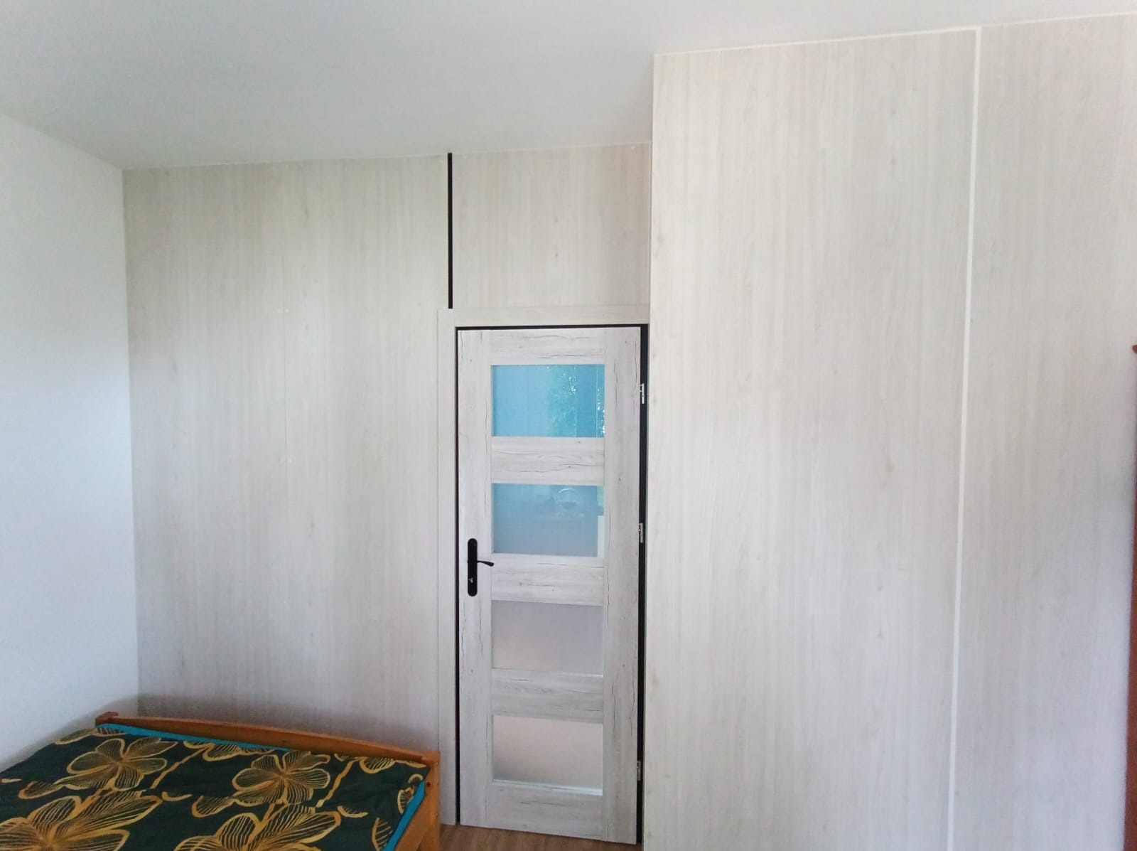 Biała szafa wnękowa z drzwiami z mlecznego szkła, widok na fragment łóżka z wzorzystą narzutą.