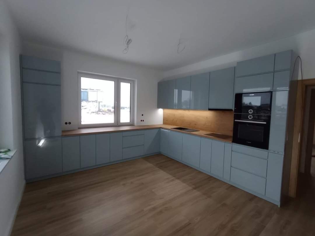 Nowoczesna kuchnia z błękitnymi, lakierowanymi frontami, drewnianym blatem i podświetleniem LED pod szafkami wiszącymi, widok na okno z placem budowy w tle.