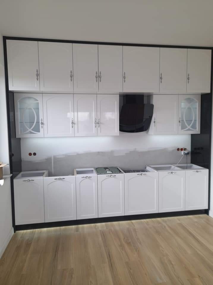 Biała zabudowa kuchenna z czarną ramą, górne szafki z przeszklonymi witrynami, widoczne niedokończone prace wykończeniowe na ścianie.