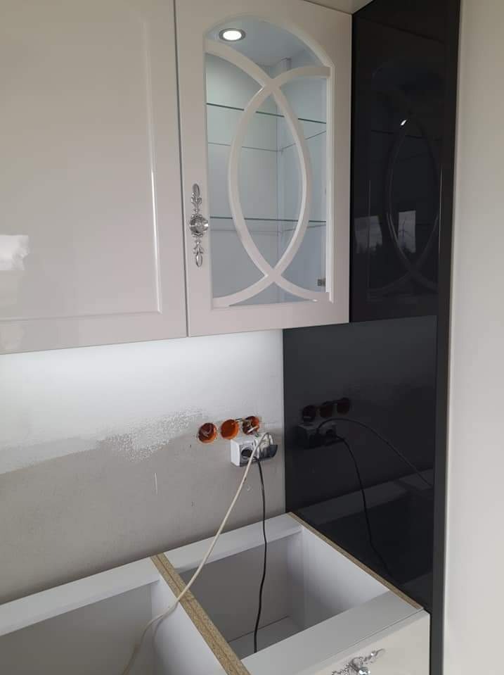 Montaż białych szafek kuchennych z oświetleniem LED i szklanymi drzwiczkami, widoczne gniazdka elektryczne i czarny panel ścienny w trakcie instalacji.