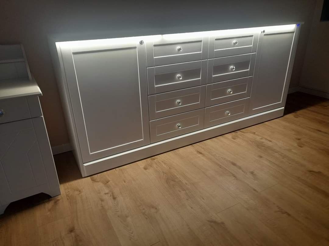 Biała komoda z szufladami i szafkami oświetlona od góry, z drewnianą podłogą w tle, wbudowana w ścianę.