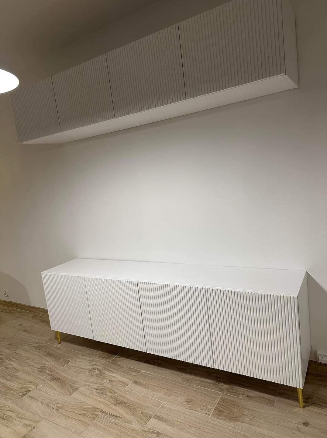 Nowoczesna, biała szafka RTV z ryflowanymi frontami i złotymi nóżkami, wisząca szafka nad nią, minimalistyczne wnętrze.