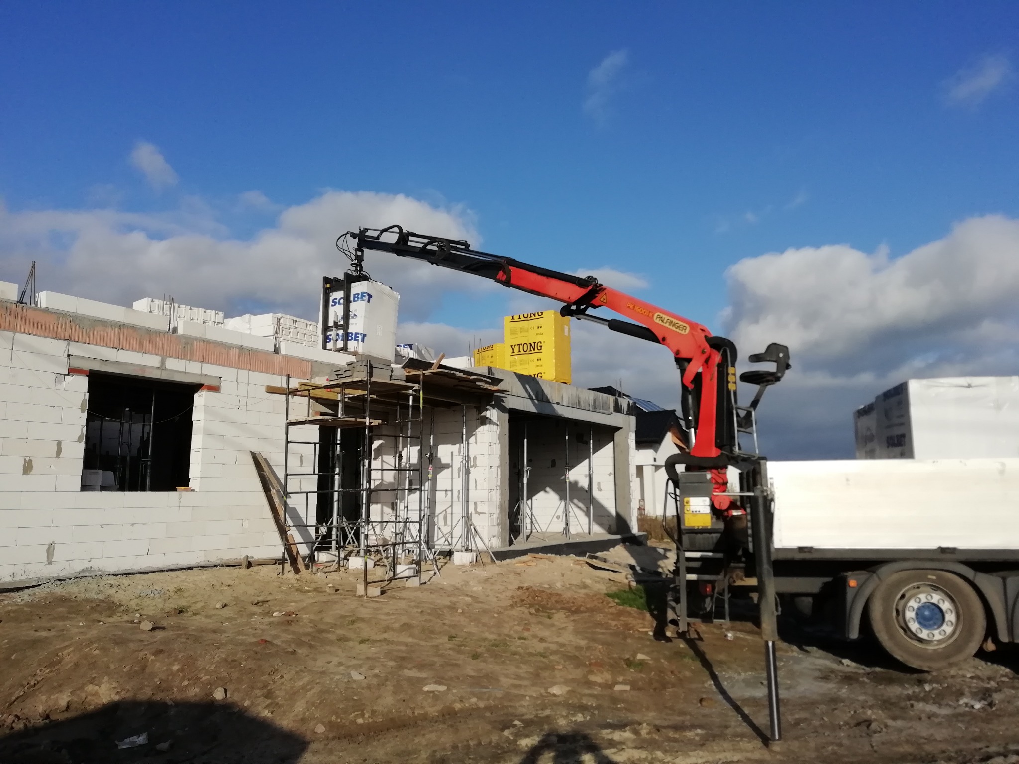 Czerwony dźwig samochodowy Palfinger podnosi pakiet białych bloczków SOLBET na budowie domu z białych bloczków, widoczne rusztowania i żółte opakowania YTONG.
