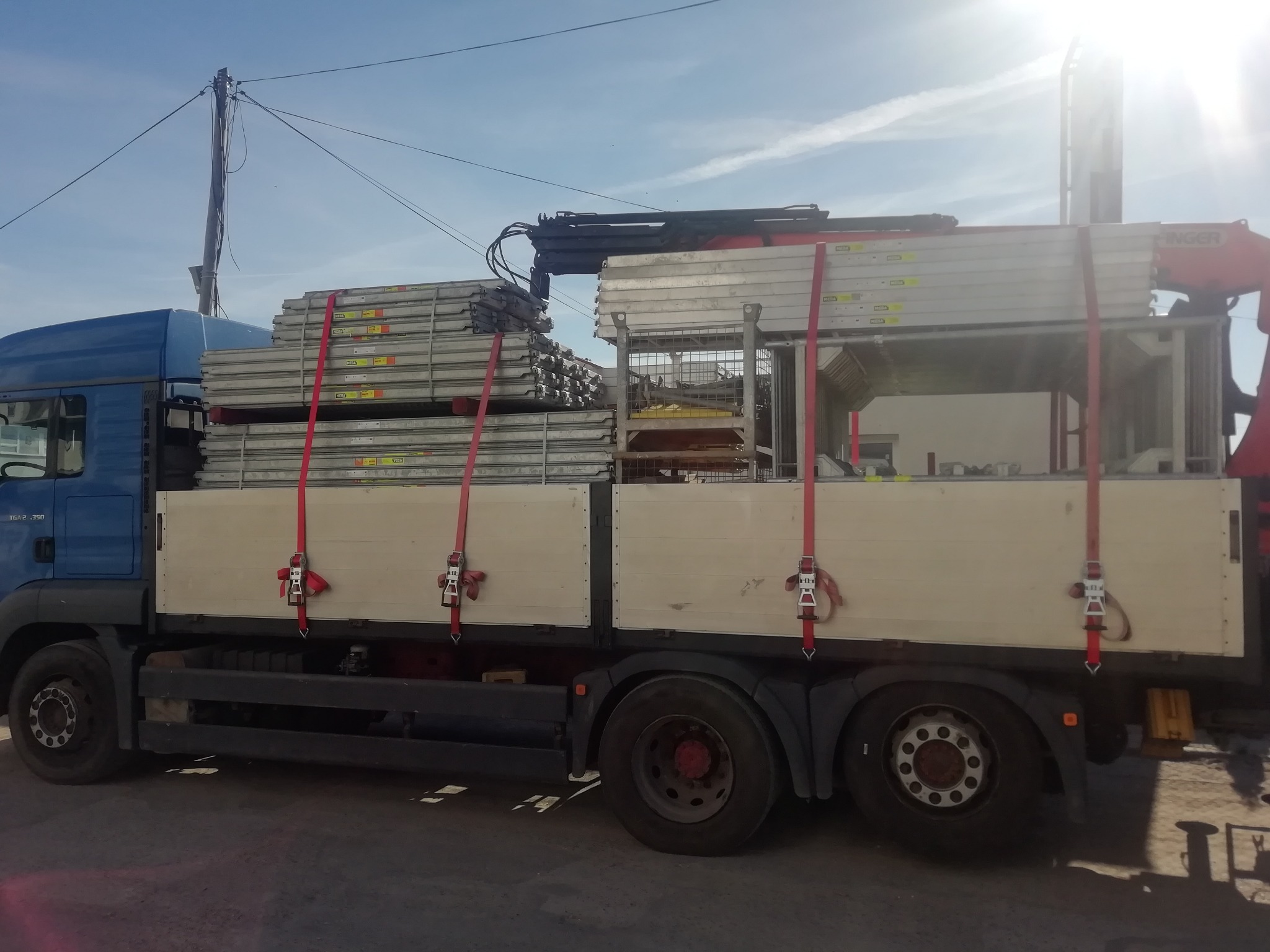 Niebieska ciężarówka MAN z platformą załadowaną metalowymi rusztowaniami zabezpieczonymi czerwonymi pasami, widoczny dźwig HMF i słup elektryczny w tle.