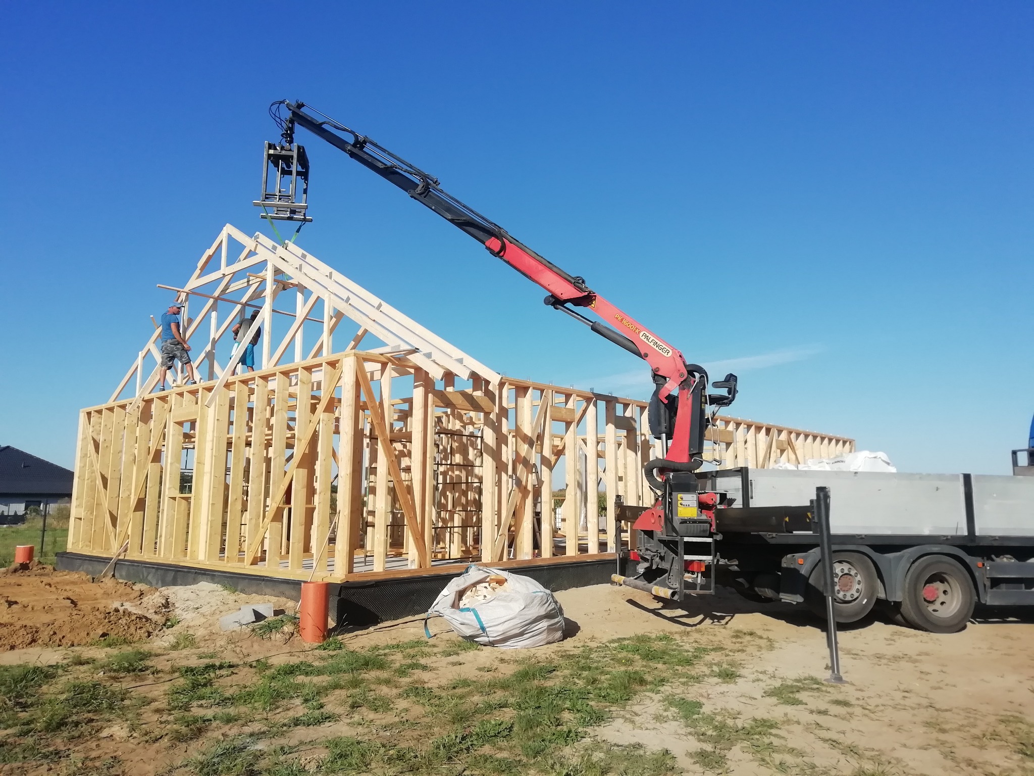 Montaż drewnianej konstrukcji domu szkieletowego z użyciem dźwigu samochodowego Palfinger na tle błękitnego nieba.