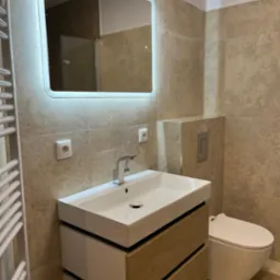 Nowoczesna łazienka z beżowymi płytkami, umywalką nablatową z szafką z drewnianymi frontami, podświetlanym lustrem i wiszącą toaletą.