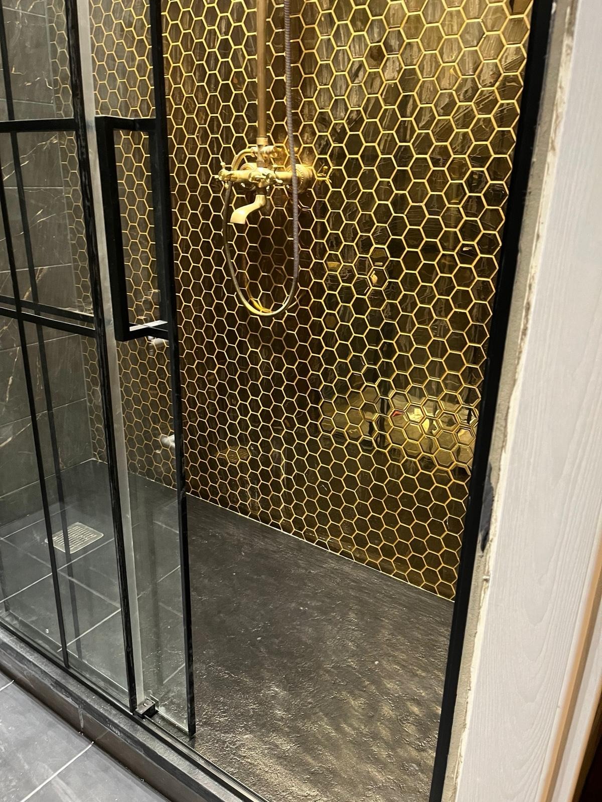 Kabina prysznicowa z czarną ramą i złotymi heksagonalnymi płytkami na ścianie, widoczna armatura w stylu retro.