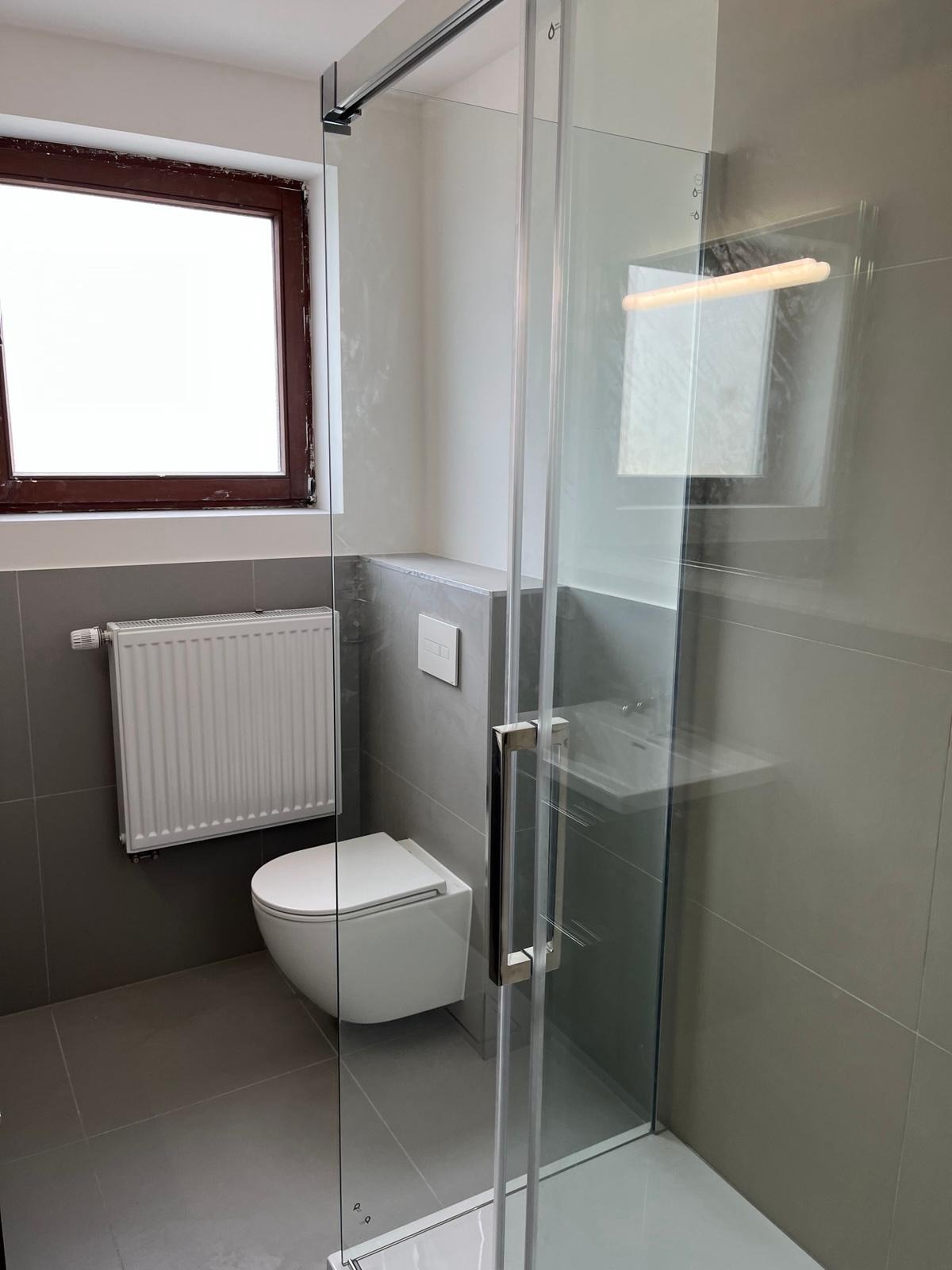 Nowoczesna łazienka z szarą glazurą, przeszkloną kabiną prysznicową, podwieszaną toaletą i grzejnikiem drabinkowym, oświetlona naturalnym światłem z okna.
