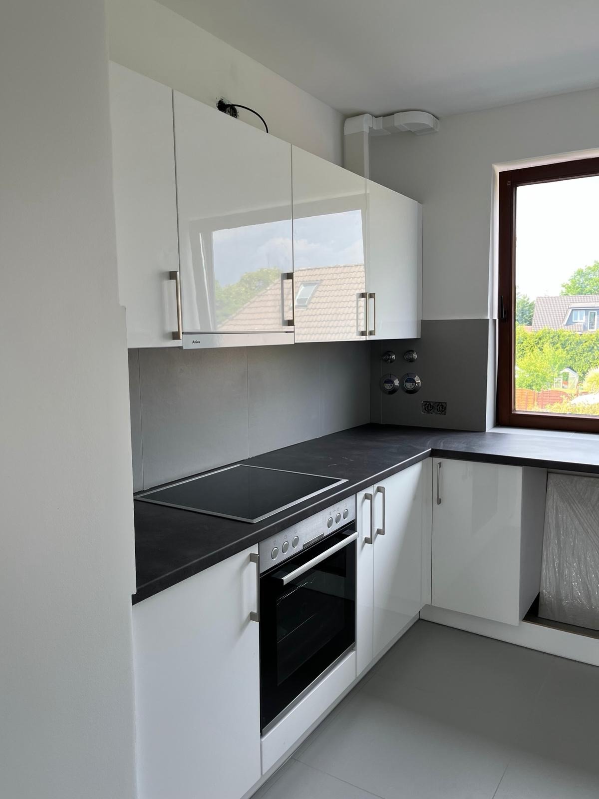 Nowoczesna kuchnia z białymi, lakierowanymi szafkami, czarnym blatem i płytą indukcyjną, widok na okno z brązową ramą i fragmentem zieleni na zewnątrz.