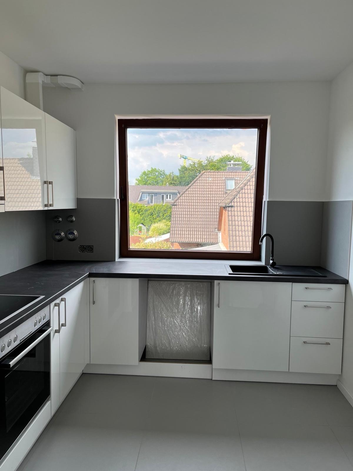 Nowoczesna, jasna kuchnia z białymi, błyszczącymi szafkami, czarnym blatem i zlewozmywakiem. Okno z widokiem na zabudowę jednorodzinną i zielone drzewa. Zmywarka oczekuje na montaż, zabezpieczona...