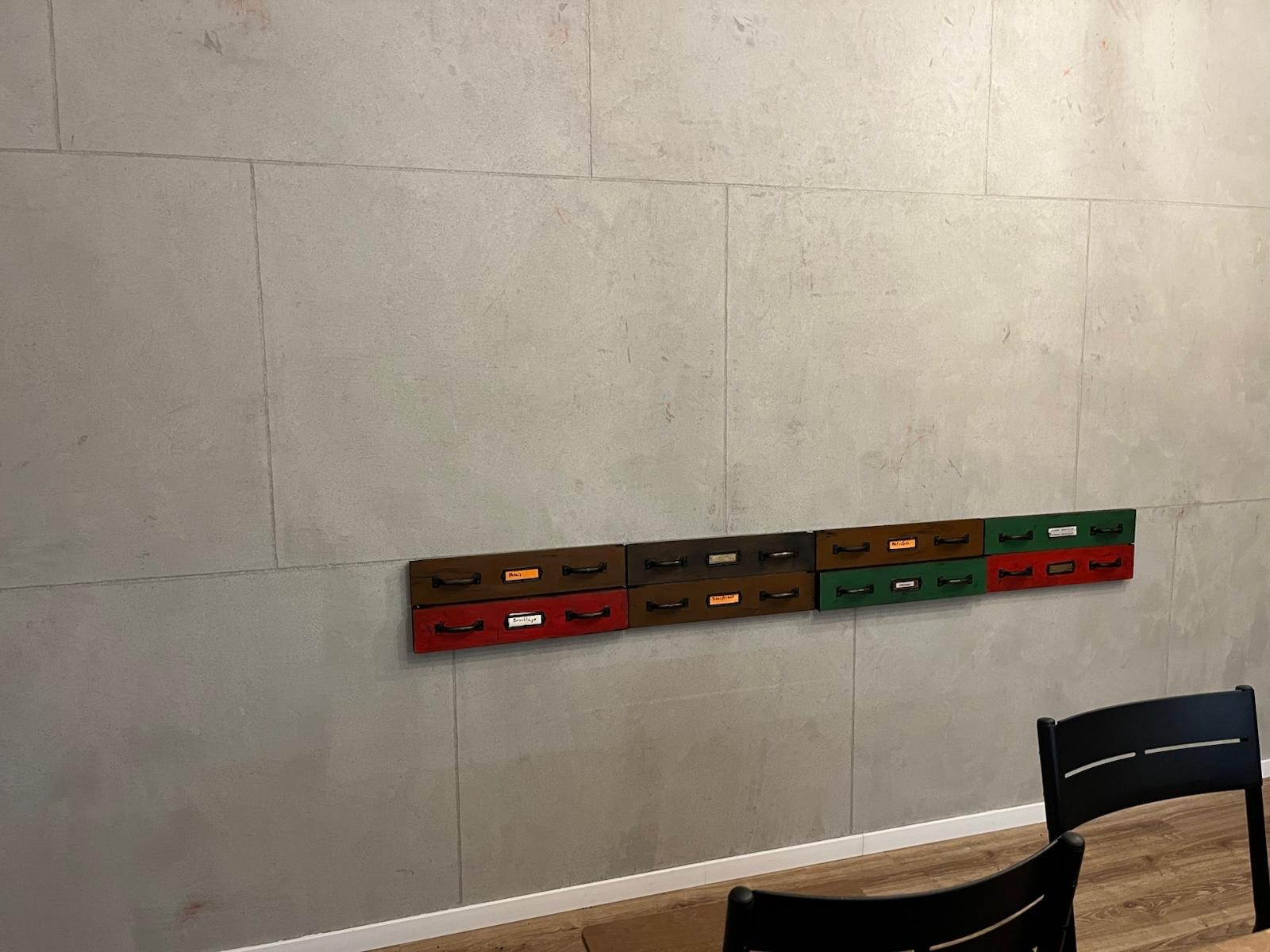 Dekoracyjna ściana z betonową fakturą i rzędem kolorowych, drewnianych szufladek z etykietami, imitujących dawne meble apteczne lub warsztatowe.