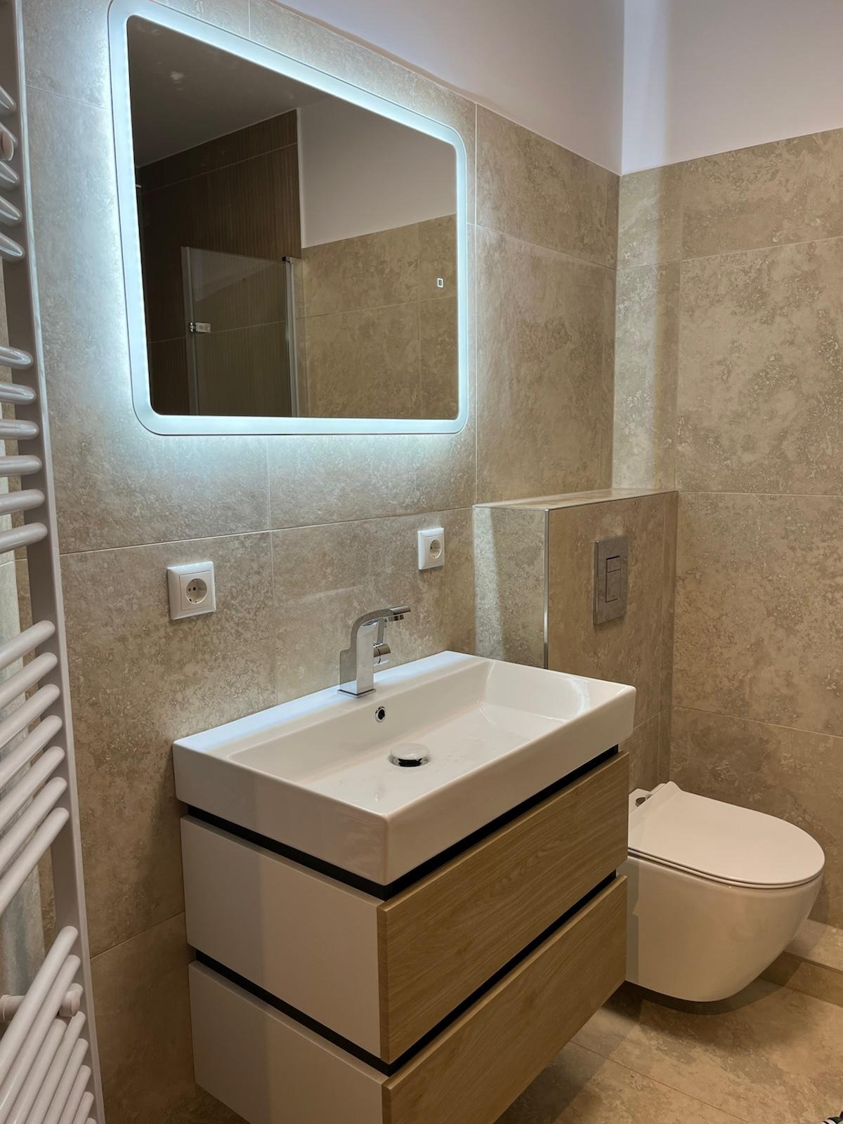 Nowoczesna łazienka z beżowymi płytkami, umywalką nablatową z szafką z drewnianymi frontami, podświetlanym lustrem i wiszącą toaletą.