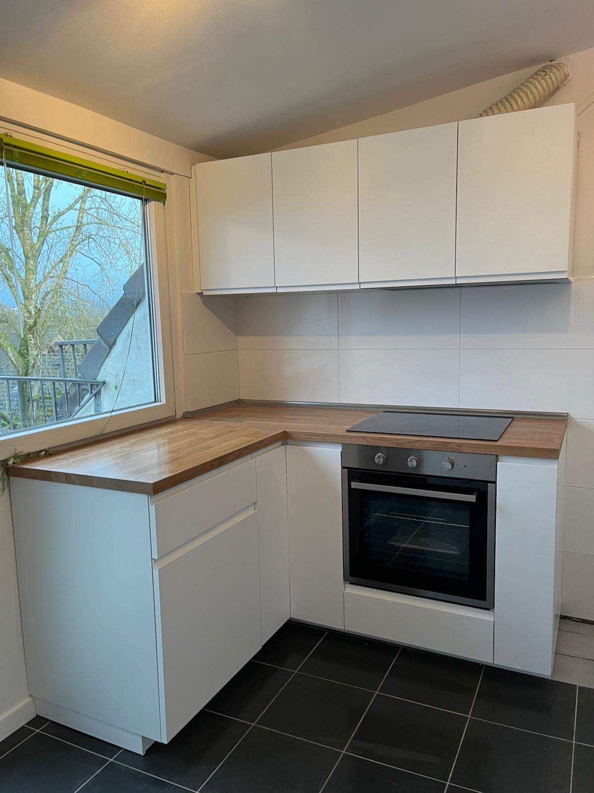 Nowoczesna kuchnia z białymi szafkami, drewnianym blatem i czarną płytą indukcyjną, widok z okna na zewnątrz.