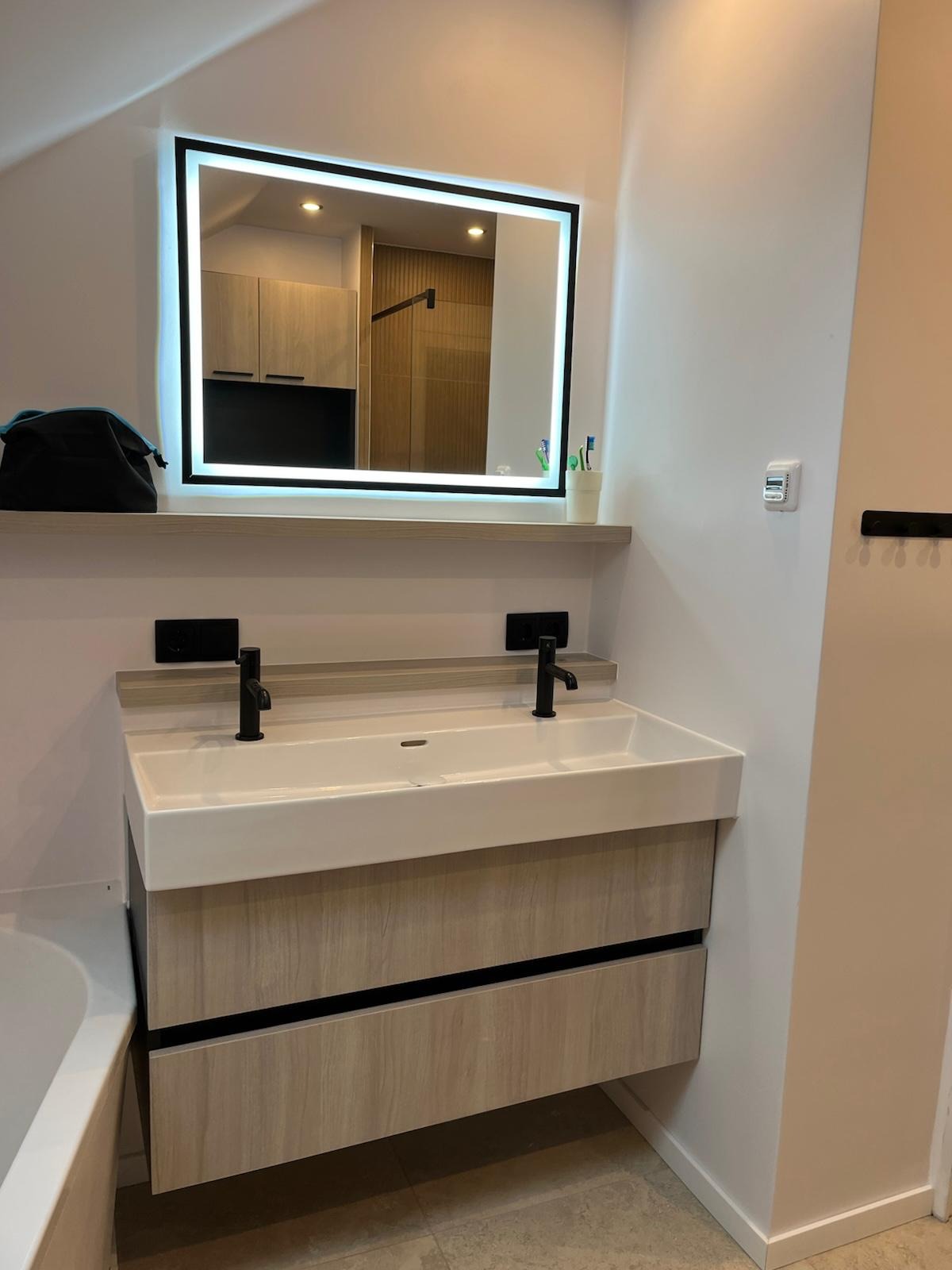 Nowoczesna łazienka z podwójną umywalką, szafką z drewnopodobnym dekorem i podświetlanym lustrem z czarną ramą, widok z boku na wannę.