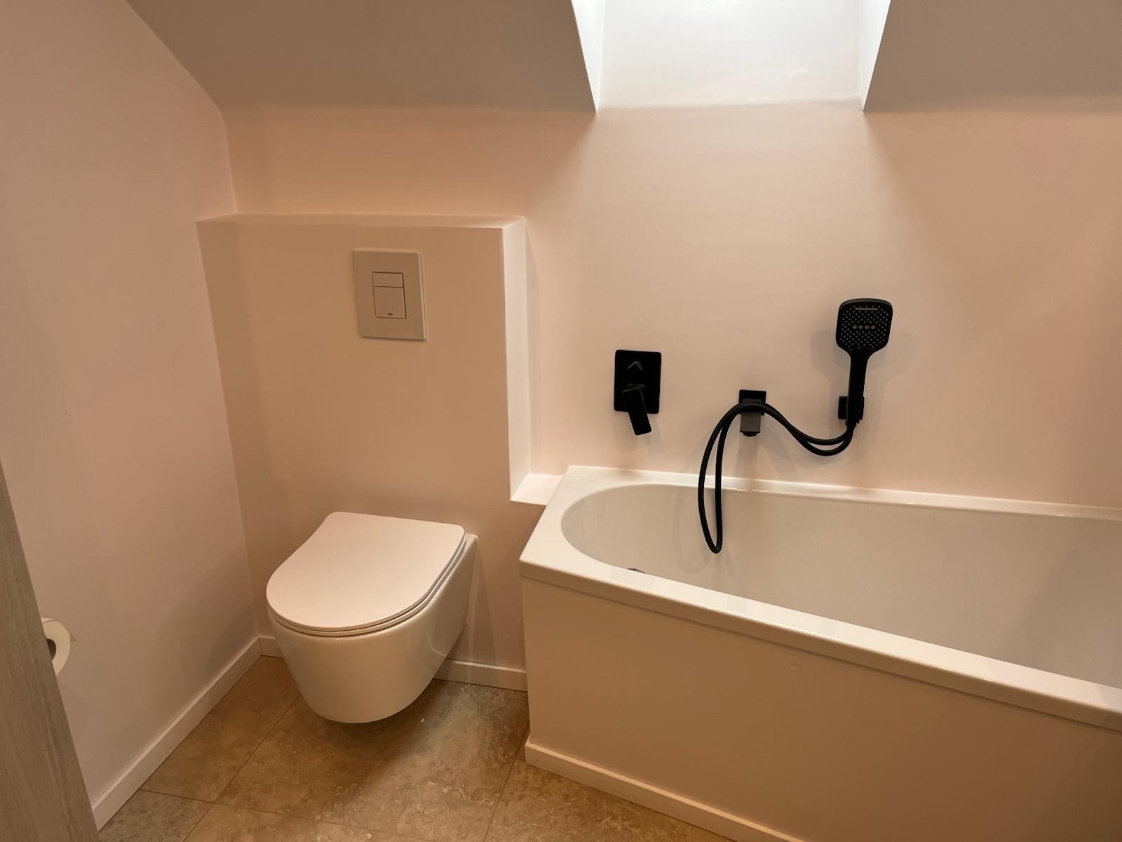 Nowoczesna łazienka z podwieszaną toaletą, białą wanną i czarną armaturą pod skosem dachu.