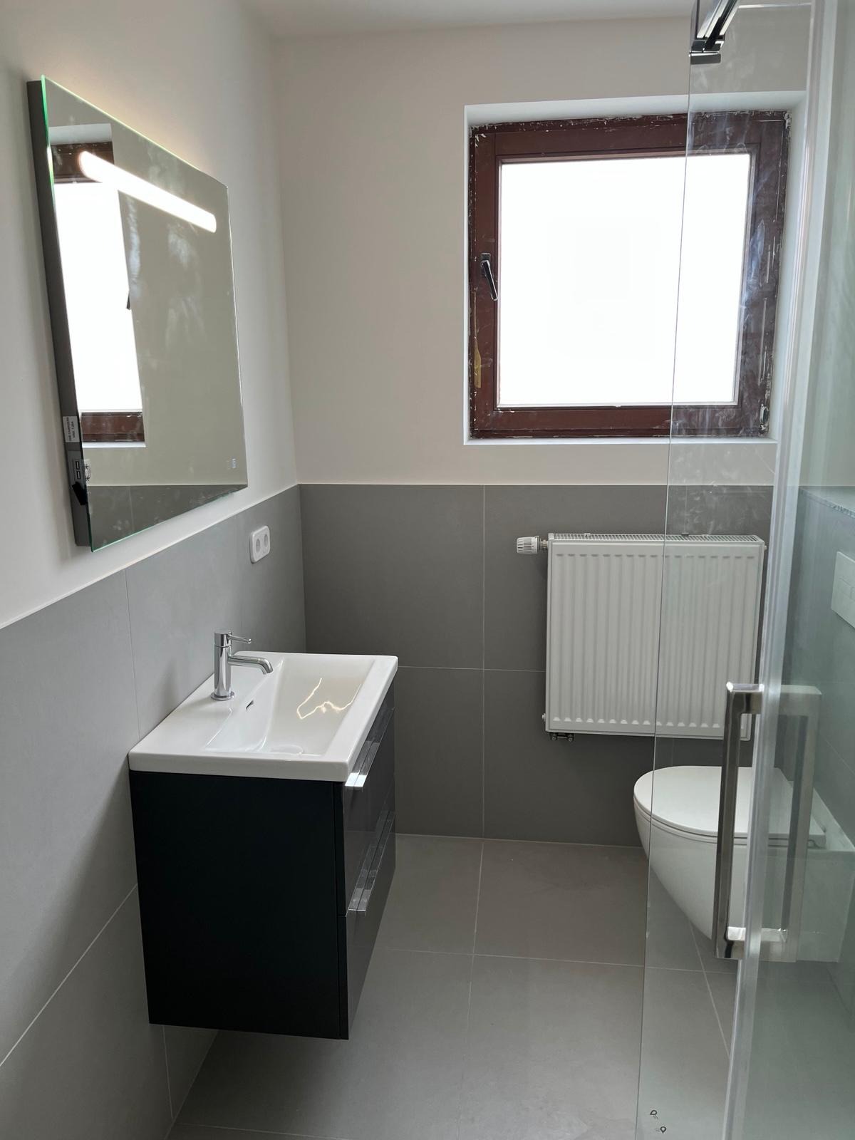 Nowoczesna łazienka z umywalką nablatową, podświetlanym lustrem, grzejnikiem panelowym i kabiną prysznicową ze szklanymi drzwiami, wykończona szarą glazurą.