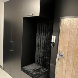 Frans meble - Ciemna zabudowa wnękowa z siedziskiem tapicerowanym czarnym welurem, obok drzwi zabezpieczone folią malarską, widoczny domofon na ścianie.