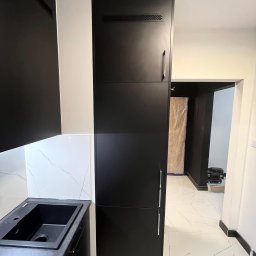 Frans meble - Czarna zabudowa kuchenna z wysoką szafą w Katowicach, widoczny zlew i fragment blatu, białe płytki imitujące marmur na ścianie, podłoga z białych płytek z szarymi żyłkami, w tle widoczne przejście...