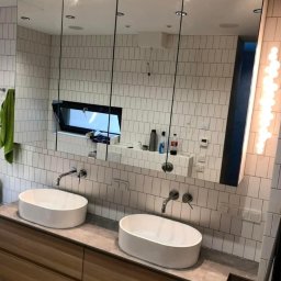 Frans meble - Łazienka z dwiema owalnymi umywalkami nablatowymi na drewnianej szafce, lustro z szafkami wbudowane w ścianę z białych płytek imitujących cegłę, okno w lustrze, oświetlenie boczne.