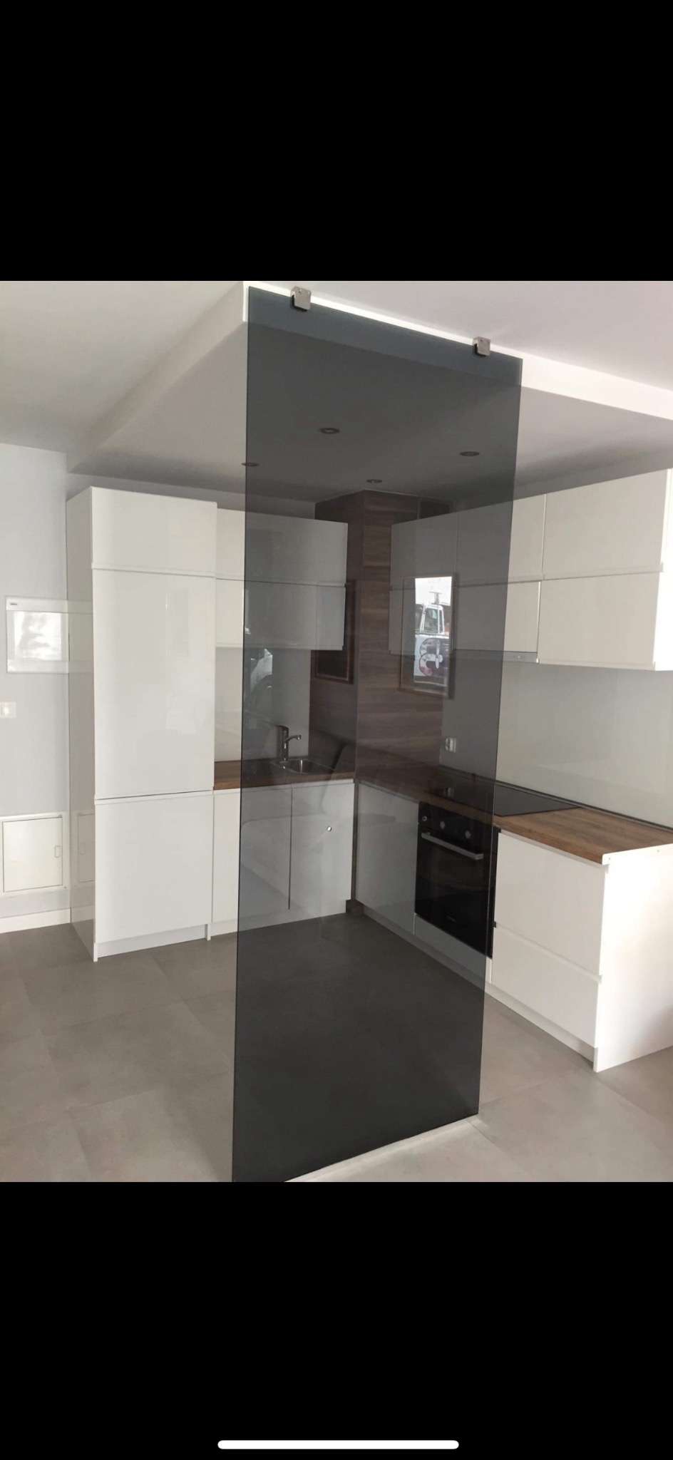 Biała kuchnia z drewnianym blatem, częściowo przesłonięta szklaną ścianką działową na szynie.