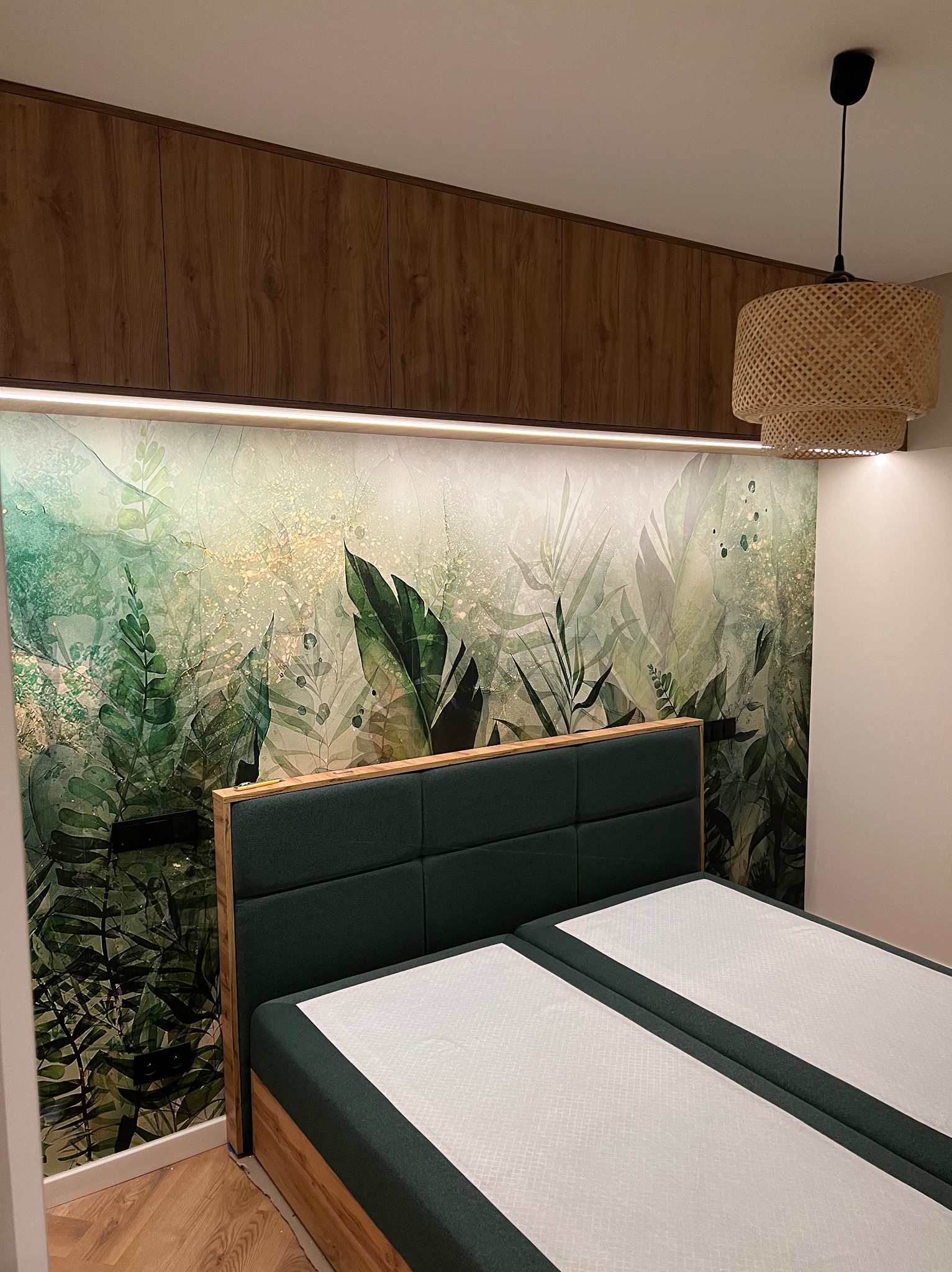 Nowoczesna sypialnia z łóżkiem dwuosobowym z zielonym zagłówkiem, szafkami wiszącymi z drewnianymi frontami i tapetą w motywy roślinne, oświetlona lampą z abażurem z naturalnego materiału.