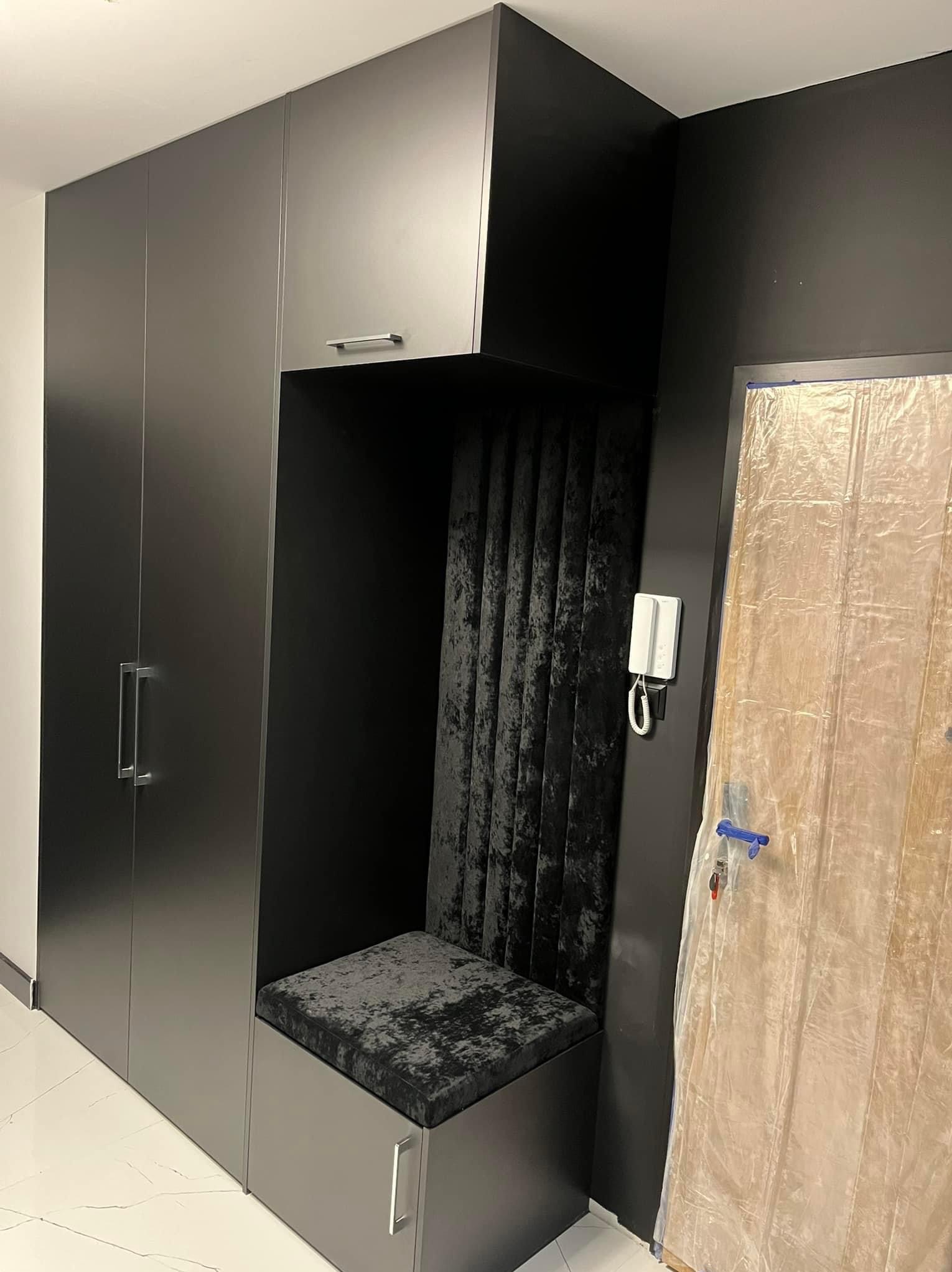 Ciemna zabudowa wnękowa z siedziskiem tapicerowanym czarnym welurem, obok drzwi zabezpieczone folią malarską, widoczny domofon na ścianie.