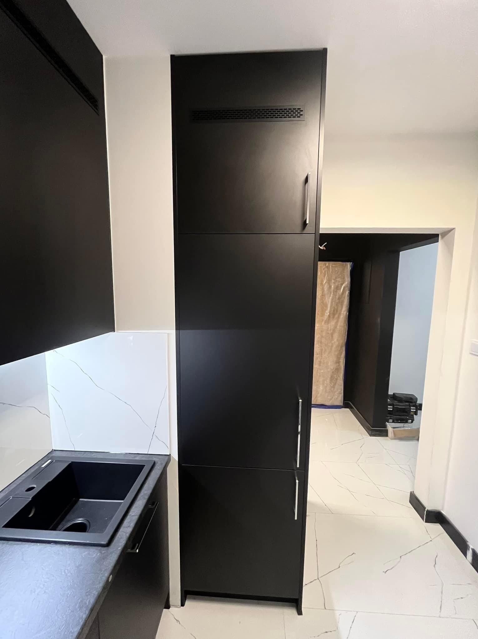 Czarna zabudowa kuchenna z wysoką szafą w Katowicach, widoczny zlew i fragment blatu, białe płytki imitujące marmur na ścianie, podłoga z białych płytek z szarymi żyłkami, w tle widoczne przejście...