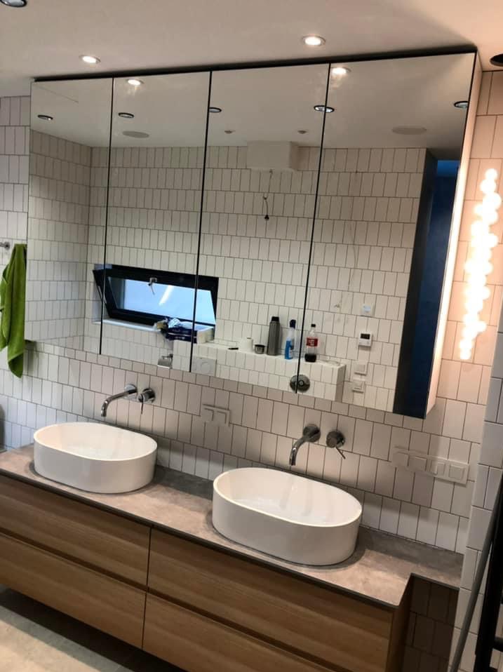 Łazienka z dwiema owalnymi umywalkami nablatowymi na drewnianej szafce, lustro z szafkami wbudowane w ścianę z białych płytek imitujących cegłę, okno w lustrze, oświetlenie boczne.