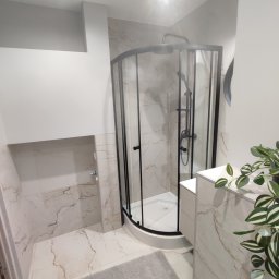 JusMar - Nowoczesna łazienka z kabiną prysznicową narożną z czarnymi profilami, wykończona płytkami imitującymi marmur, uzupełniona szarą wycieraczką i rośliną doniczkową.