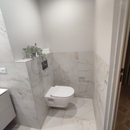JusMar - Nowoczesna toaleta podwieszana w łazience wykończonej płytkami imitującymi marmur, z akcentami dekoracyjnymi w postaci rośliny w doniczce i minimalistycznego włącznika światła.