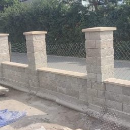 JusMar - Wznoszony mur z bloczków betonowych z filarami i podmurówką, widoczne narzędzia i materiały budowlane na placu budowy, ogrodzenie z siatki w tle.