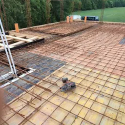 Zbrojenie stropu budynku w trakcie budowy, widoczne stalowe pręty tworzące siatkę, szalunki z desek, wystające pionowe pręty zbrojeniowe, narzędzie do wiązania zbrojeń i fragment butów pracownika...