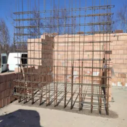 Zbrojenie pionowe i poziome ścian z bloczków ceramicznych, przygotowane do zalania betonem, widok na plac budowy z samochodem dostawczym w tle.