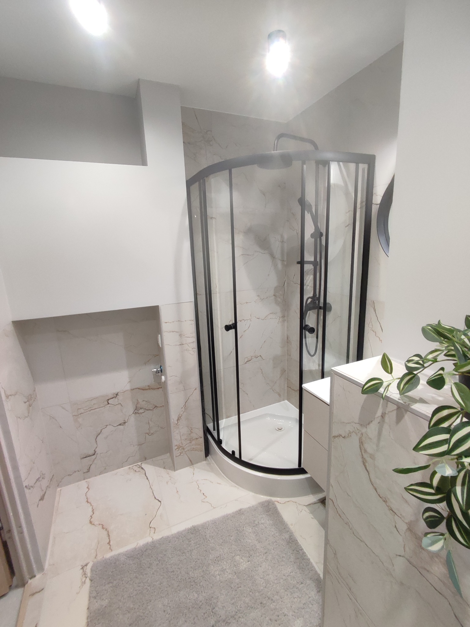 Nowoczesna łazienka z kabiną prysznicową narożną z czarnymi profilami, wykończona płytkami imitującymi marmur, uzupełniona szarą wycieraczką i rośliną doniczkową.
