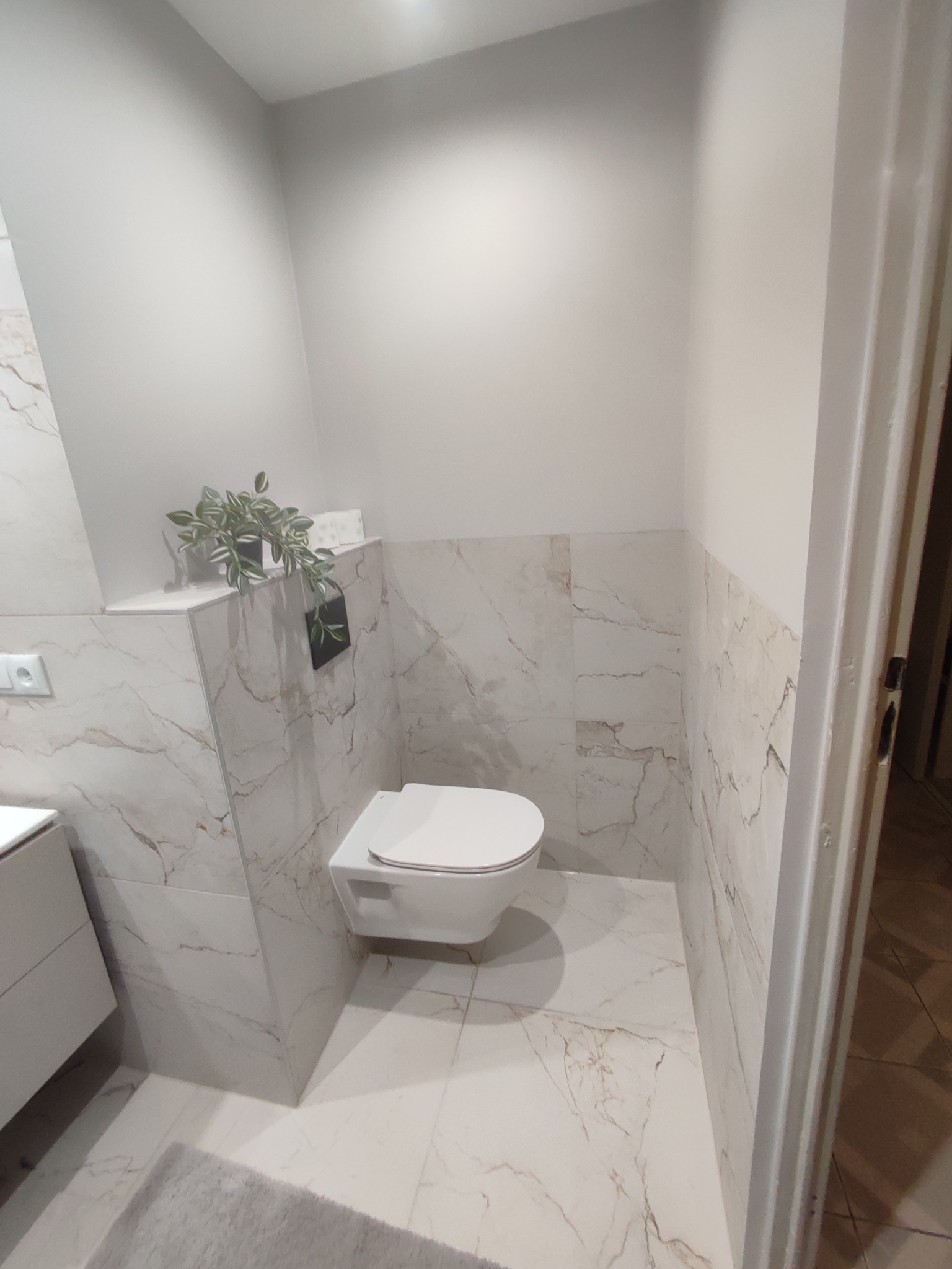 Nowoczesna toaleta podwieszana w łazience wykończonej płytkami imitującymi marmur, z akcentami dekoracyjnymi w postaci rośliny w doniczce i minimalistycznego włącznika światła.