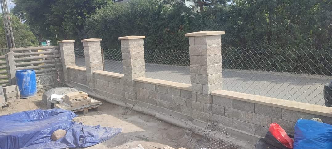 Wznoszony mur z bloczków betonowych z filarami i podmurówką, widoczne narzędzia i materiały budowlane na placu budowy, ogrodzenie z siatki w tle.