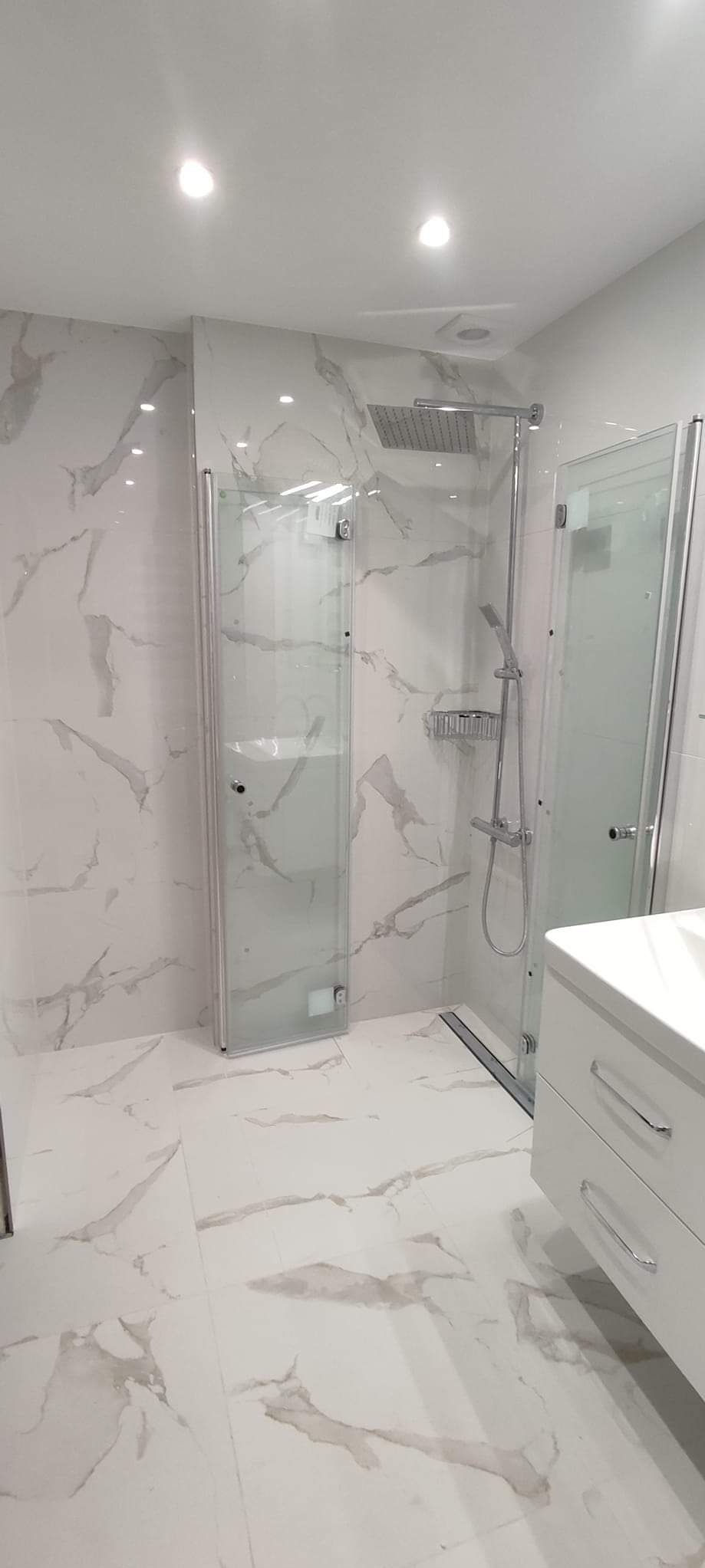 Nowoczesna łazienka z kabiną prysznicową typu walk-in, wykończona białymi płytkami imitującymi marmur, z chromowaną armaturą i białą szafką pod umywalkę z szufladami.