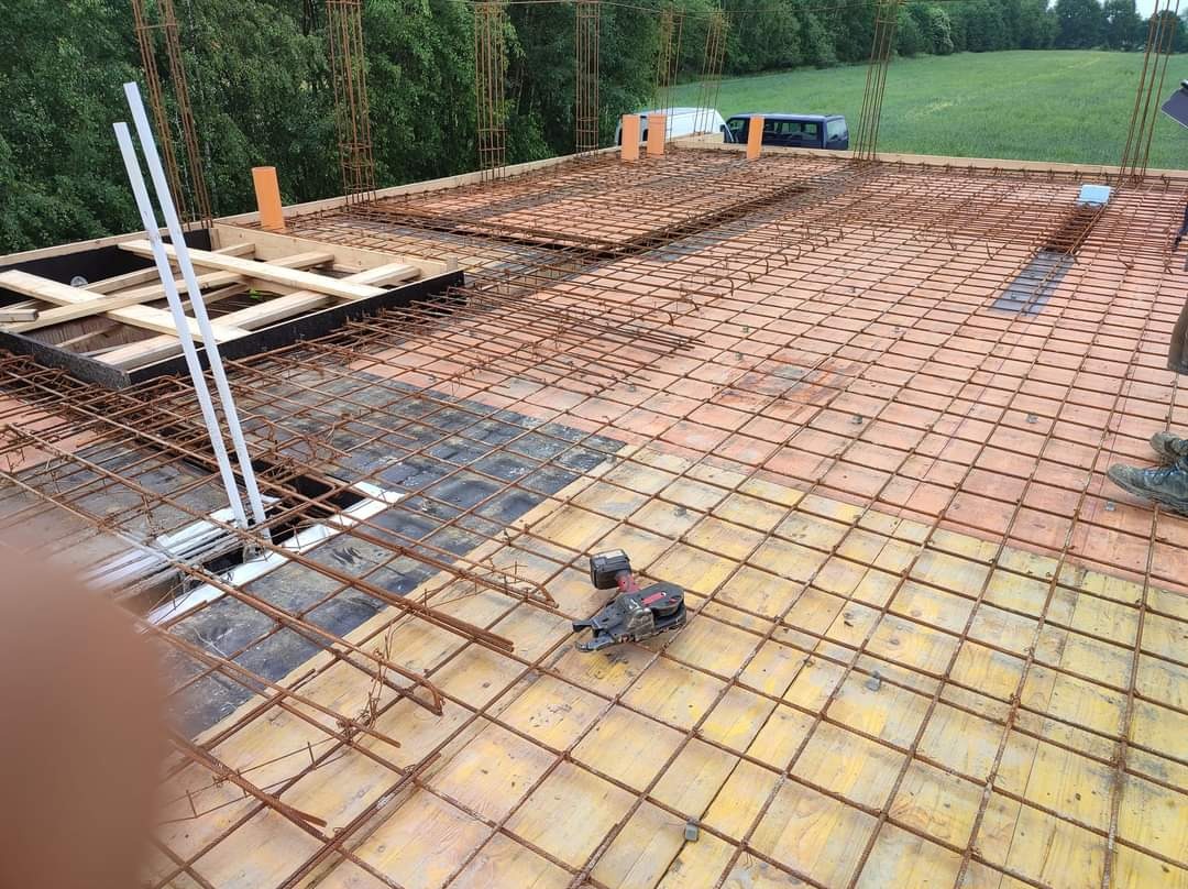 Zbrojenie stropu budynku w trakcie budowy, widoczne stalowe pręty tworzące siatkę, szalunki z desek, wystające pionowe pręty zbrojeniowe, narzędzie do wiązania zbrojeń i fragment butów pracownika...