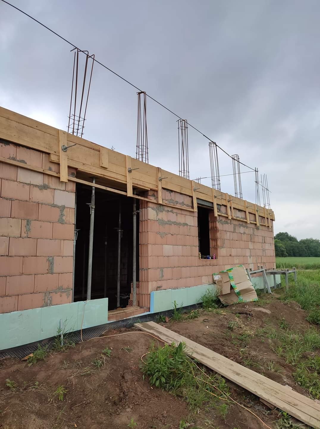 Wznoszenie murów domu z czerwonej cegły, widoczne szalunki i wystające pręty zbrojeniowe, ocieplenie fundamentów styropianem, teren budowy z ziemią i trawą.